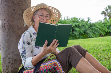 Portrait of relaxed attractive senior woman with hat and eyeglasses sitting on the meadow of public park reading a book - caucasian lady enjoying free time and retirement