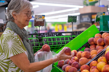 Senior woman wearing protective gloves selecting fresh peaches in the supermarket - consumerism concept, rising prices, inflation