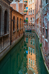 Reflections of the sky and houses on the surface of the water of a narrow canal street in Venice, old walls of houses with windows on a Venetian street, a lonely boat near the brick wall of the house