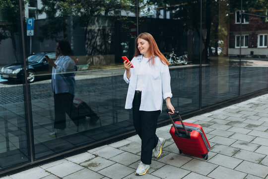 Business Woman with a red suitcase using smartphone in the European city. Hipster girl browsing Internet on a phone and waiting taxi and communicating outdoors. Mobile Travel Maps.