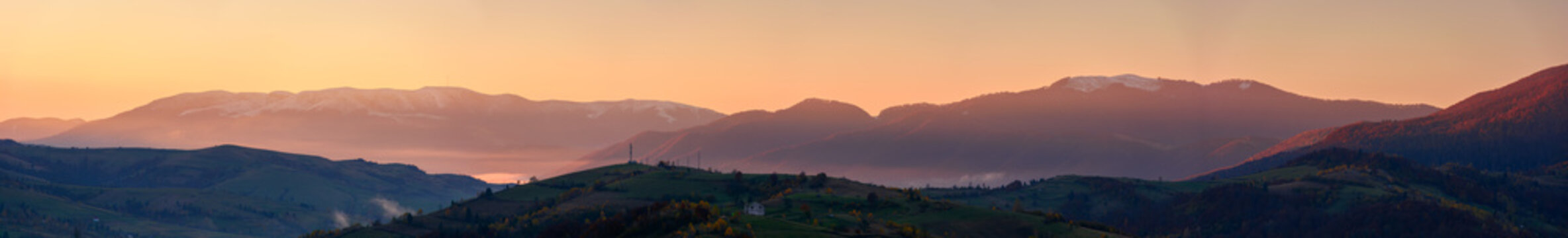 panorama of mountainous countryside at dawn. hills with trees and rural fields rolling down into the foggy valley. distant ridge with snow capped tops beneath a sky with clouds in red morning light