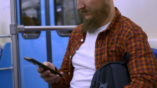A man with a beard sits in a subway car and has fun on the Internet using a smartphone. Pass the time while traveling in the meter in social networks using a smartphone.