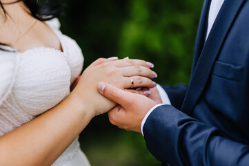 The groom in a blue suit and the bride in a white dress stand at the ceremony holding hands with a gold ring on their finger. Wedding photography close up, portrait, macro.