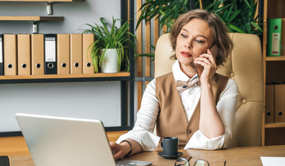 Confident stylish mature middle aged elderly woman in office.Workplace with laptop,using talking by...
