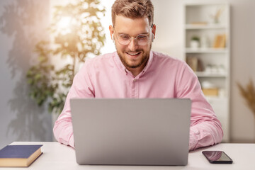 Fototapeta premium Happy handsome bearded man in eyewear working on laptop, smiling male work from home, light interior of living room