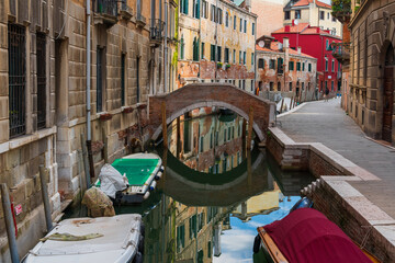 Reflections of the sky and houses on the water surface of a narrow canal street in Venice, boats, old walls of houses with windows on a Venetian street, a stone bridge over a canal
