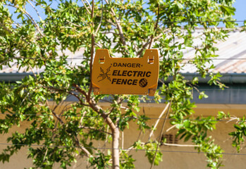 Electric fence and yellow warning sign, near a building in African town. Wire fence with cable of high voltage for safety from criminality and wild animals in urban infrastructure.