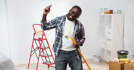 Happy African American handsome young guy dancing with paint brush roller in hand listening to...