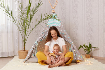 Indoor shot of happy woman wearing casual style sitting on floor near wigwam with her baby daughter, playing and having fun, spending time together. Motherhood and childhood. © sementsova321