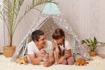 Horizontal shot of brunette Caucasian handsome man wearing white t-shirt with his child posing on floor near wigwam and using cell phone, watching cartoons or funny videos together. © sementsova321