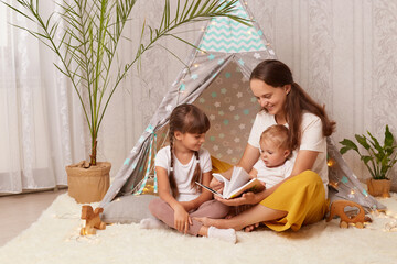 Indoor shot of mother with two charming little girls sitting together in wigwam, mom with her children reading interesting book with fairy tale in peetee tent. © sementsova321