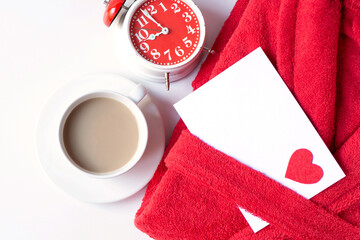 A terry red bathrobe, a cup of coffee latte, an alarm clock and a love letter are on a white...