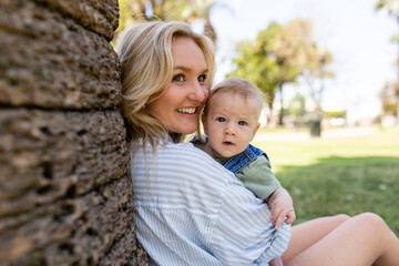 Young Mother Leans Against A Tree In The Park While Holding Her Baby