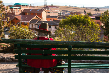 Obraz premium Old woman, in typical Peruvian dress, sitting with her back on a bench in a park with a view of the city of Chinchero, Peru. 