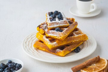 Stacked pumpkin waffles, sprinkled with powdered sugar, with fresh blueberries on a white plate on a light concrete background. Waffle recipes. Pumpkin dishes. Thanksgiving Day