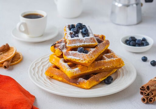 Stacked pumpkin waffles, sprinkled with powdered sugar, with fresh blueberries on a white plate on a light concrete background. Waffle recipes. Pumpkin dishes. Thanksgiving Day