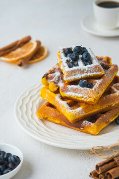 Stacked pumpkin waffles, sprinkled with powdered sugar, with fresh blueberries on a white plate on a light concrete background. Waffle recipes. Pumpkin dishes. Thanksgiving Day