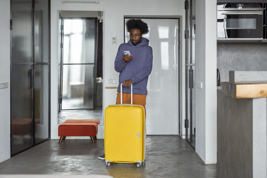 Black traveler with suitcase using cellphone at home