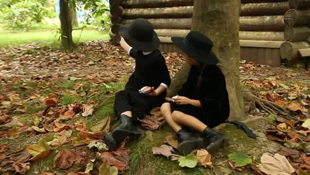 Two little girls, in black witch costumes, and hats, paint each other with scary makeup and laugh, on Halloween
