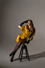Fototapeta na wymiar fashion model girl shooting in a mustard-colored dress on a white background on a cyclorama sitting on a chair subdued light 