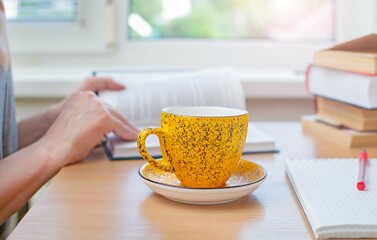 A woman in home clothes is sitting at a table with a cup of coffee and reading a printed book. Selective focus, tinting.