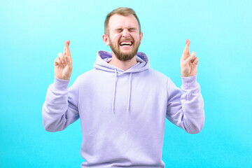 Man with a wry face crossing his fingers on a blue background. Hoping for luck. Panic. Fright. Suffer. Scared. Scary. Feared. Anxious. Frightened. Afraid. Frowning. Stress. Upset. Frustration