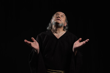 medieval monk in dark cassock looking up while praying with outstretched hands isolated on black.