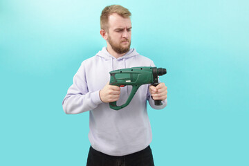 A man in a purple hoodie holding a drill and suspiciously look at it on a blue background....