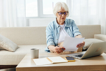 an elderly lady works sitting on the couch at home reading a text from a paper during an online conference with colleagues