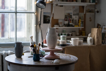 Clay vase dries up standing on wooden rack. Masterpiece prepared for making sketch and painting stands on table against tools for sculpturing. Handmade vase created for sending to exhibition in museum