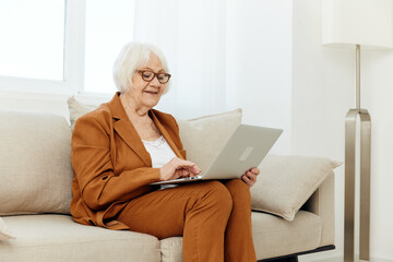 Mila is a joyful old lady sitting with a laptop on her lap on a beige sofa near the window and looking at the monitor with a pleasant smile on her face working after retirement