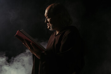 side view of senior priest in cassock reading holy bible on black with smoke.