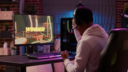 Over shoulder view of african american gamer celebrating after winning online competition on gaming pc. Man doing victory hand gesture happy after playing online multiplayer game first person shooter.