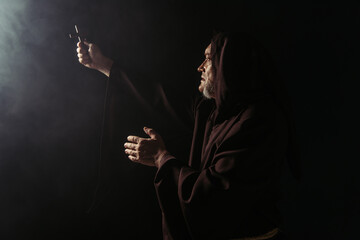 Fototapeta na wymiar monk in black hooded robe holding holy crucifix in outstretched hand on dark background.