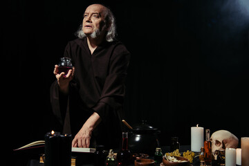 thoughtful alchemist holding jar with liquid near magic cookbook on black background.