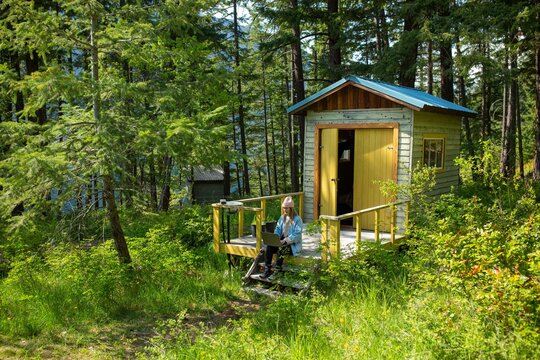 A woman is sitting outside a cabin working on her computer 
