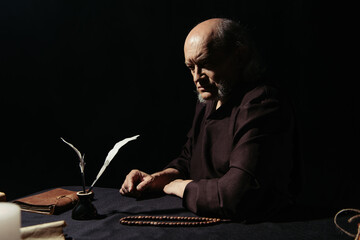 thoughtful monk sitting near inkpot and rosary at night isolated on black.