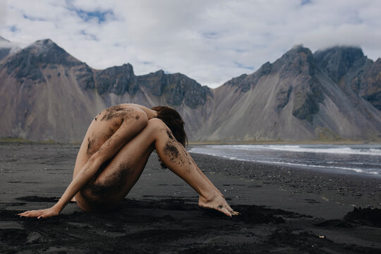 Naked woman sitting on sand near the sea in front of mountains Iceland
