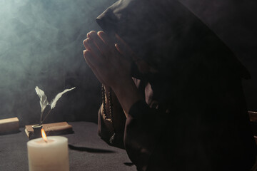 medieval monk in hooded cassock praying with rosary beads near burning candle on black background with smoke.
