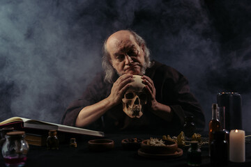 Fototapeta na wymiar discouraged alchemist holding skull near ingredients and magic cookbook on black background with smoke.