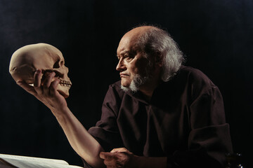 senior philosopher in dark robe looking at human skull isolated on black.