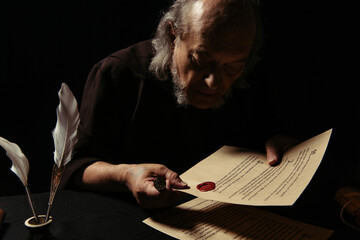 senior priest holding wax seal and looking at stamped manuscript isolated on black.