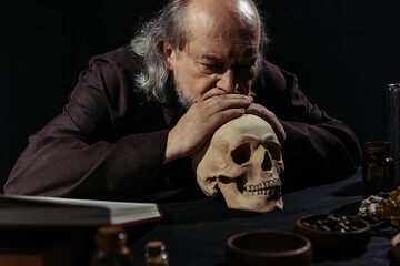 senior alchemist thinking near skull and bowls with dried herbs isolated on black.