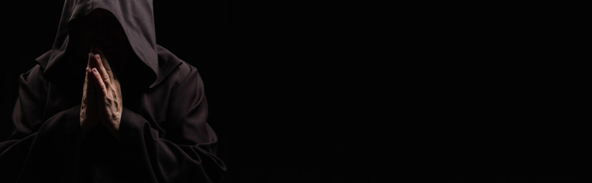 priest with face obscured by dark hood praying isolated on black, banner.