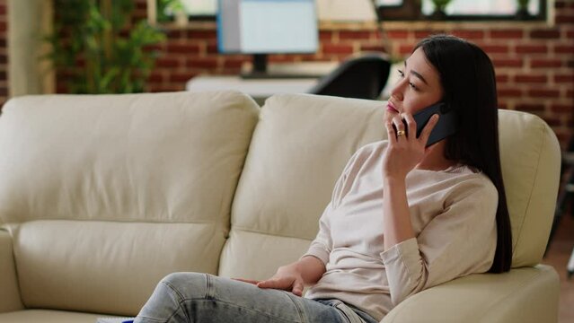 Young woman working from home while talking on smartphone sitting on couch in living room. Happy person discussing with friend on modern mobile phone while doing remote work.