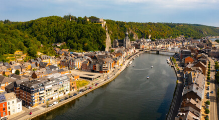 Aerial view of Walloon city of Dinant located along Meuse river with Gothic medieval Collegiate Church of Our Lady and fortified castle perched atop green vertical cliff in summer, Belgium - obrazy, fototapety, plakaty