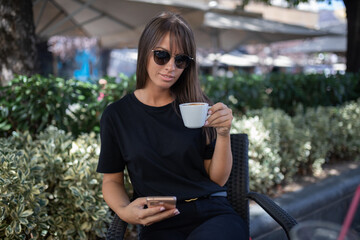 Morning in cafe - attractive woman in black dress and dark sunglasses drinkin coffe and make selfie photo for social networks