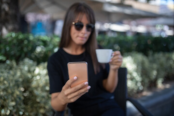 Morning in cafe - attractive woman in black dress and dark sunglasses drinkin coffe and make selfie photo for social networks