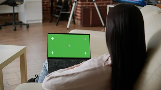 Woman sitting on couch inside apartment with portable computer having green screen display. Young adult person sitting on sofa at home while looking at laptop having chroma key isolated background.
