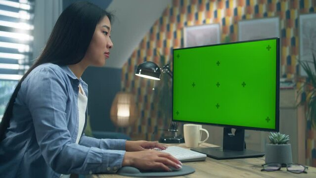 Asian woman sitting at the table and looking at screen of personal computer while working remotely from home or surfing the internet. Freelance. Green screen and chromakey